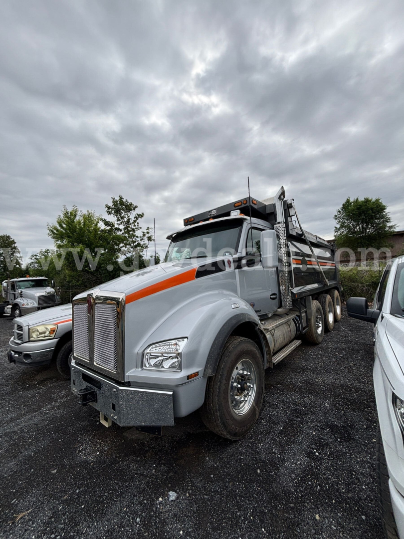Kenworth T880 dump truck in a parking lot, grey with orange accents, front grille, dual rear wheels, dump bed visible.