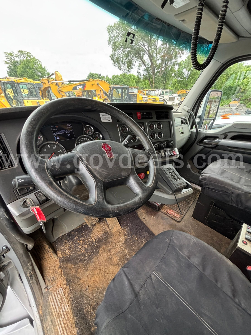 Kenworth T880 truck interior showing steering wheel, dashboard, gear shifter, and dirty floor mats; view through windshield with construction equipment outside.