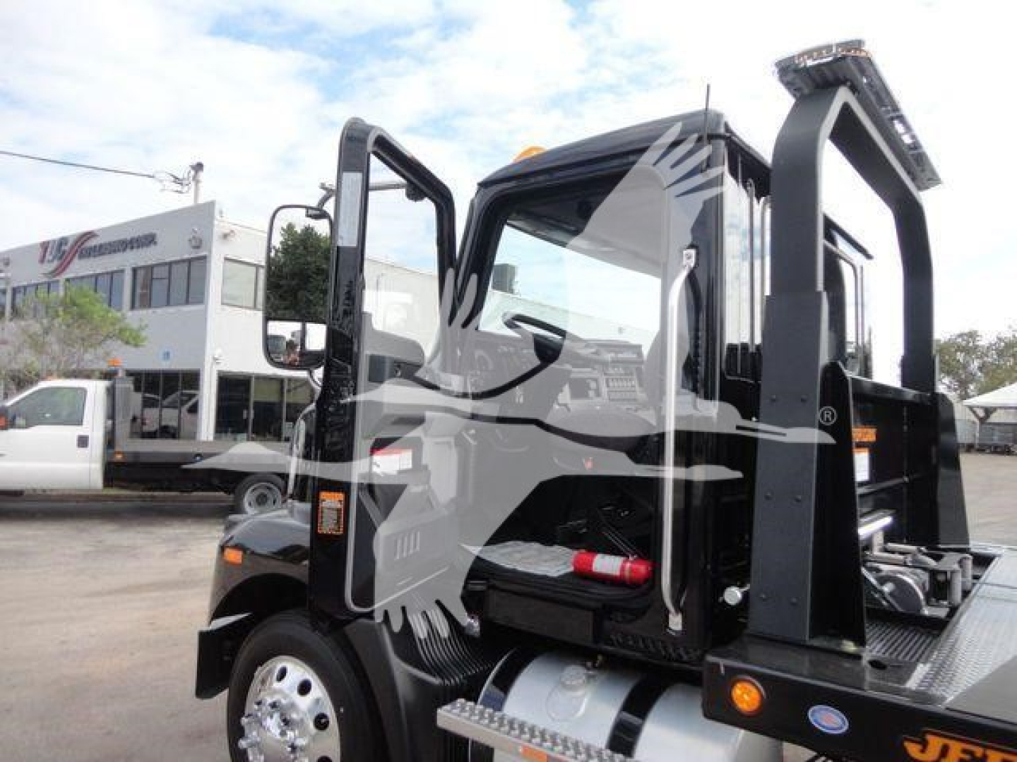 Truck cab interior/exterior of a 2027 Mack MD6 rollback truck with open driver door, black painted frame, chrome wheels, and hydraulic uplift crane visible.