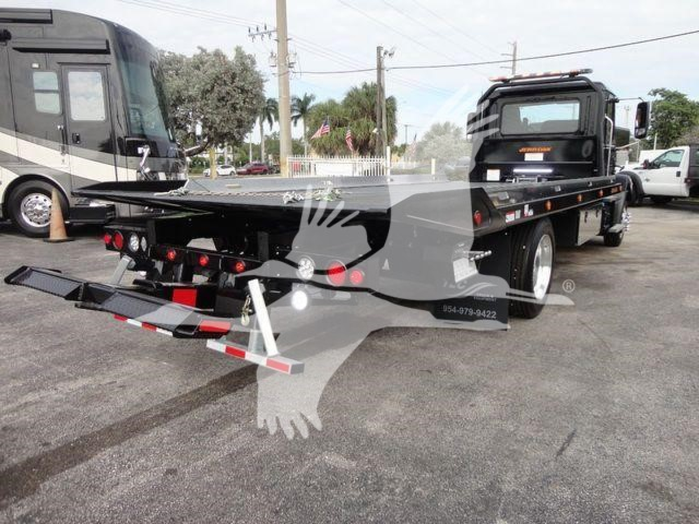 Flatbed tow truck with rollback equipment and hydraulic tilting bed; rear bumper and taillights visible; company graphics on cab; text on bumper string shows numbers.