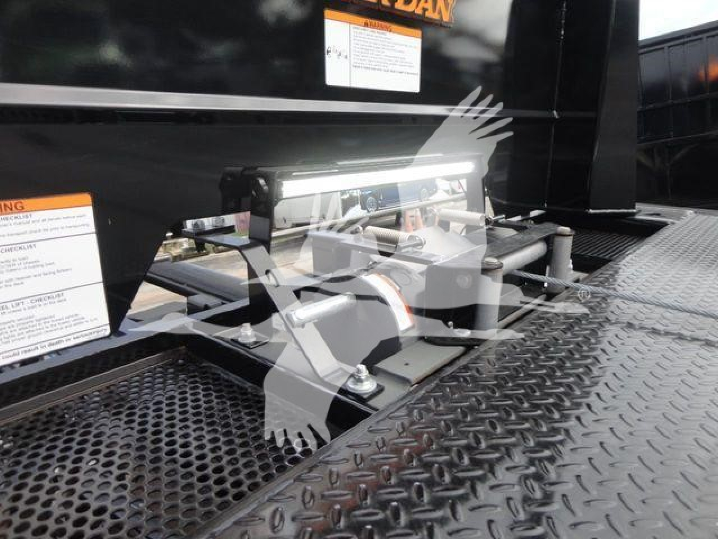 Tractor-trailer style equipment with hydraulic hose connections and mounting brackets on a diamond-plate surface; labels and warning stickers visible on black surface.
