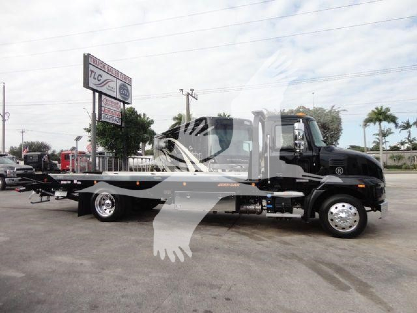 Truck with rollback equipment viewed from side, cab is black, long flatbed with hydraulic ramps, dealership lot in background.