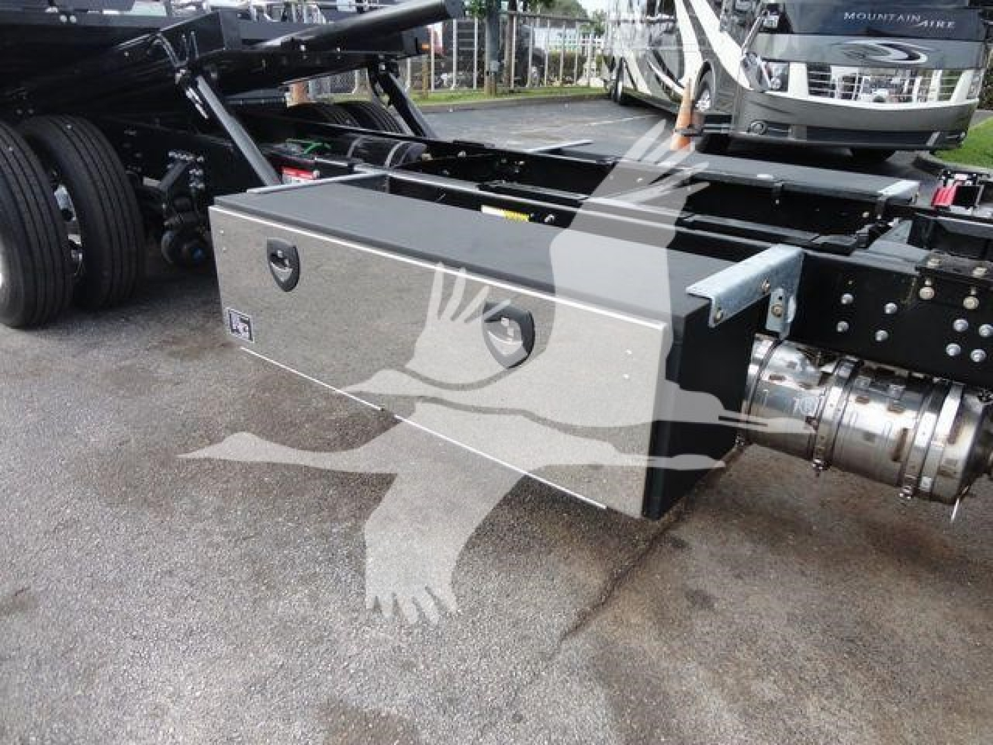 Tractor-trailer chassis with a rectangular metal toolbox mounted near the rear, side view showing two circular compartments on toolbox lid and chrome drive shaft nearby.