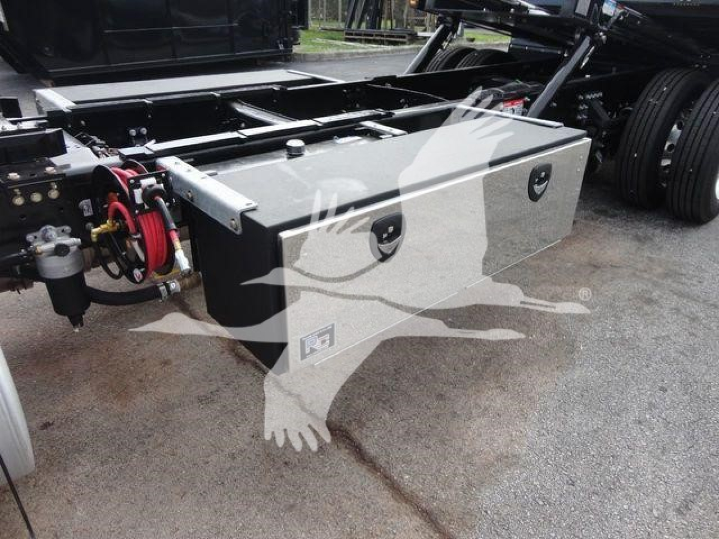 Truck toolbox mounted on frame with hydraulic components and wiring; metal storage box with two circular access ports and a small logo on front.