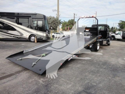 Tow truck flatbed rollback equipment on a trailer with a ramp, side rails, and winch hooks visible; MD6 truck in background.