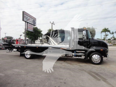 2027 Mack MD6 rollback truck on lot with black cab and long flatbed, logo signs in background.