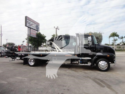 Truck with rollback equipment viewed from side, cab is black, long flatbed with hydraulic ramps, dealership lot in background.
