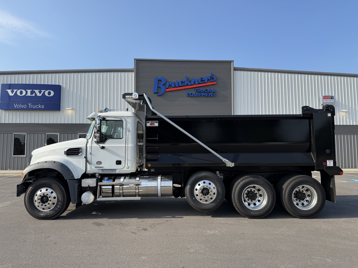 Dump truck side view with black dump bed and white cab in front of a truck dealer building. Visible text includes Bruckners Truck Equipment sign, Volvo Trucks banner, and the truck model is 2027 Mack GR84F (not visible on the image but in l
