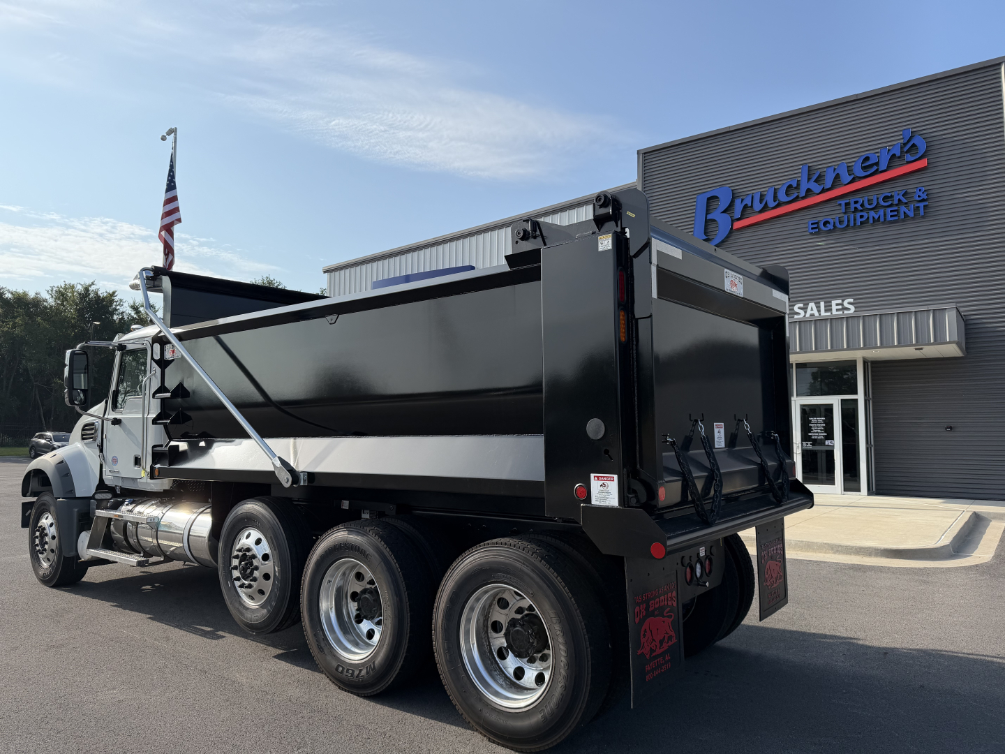 Dump truck rear and side view showing Mack GR84F dump body in black with white cab, triple rear axle configuration, ramp at tail, and Brückner’s Truck & Equipment sales sign.