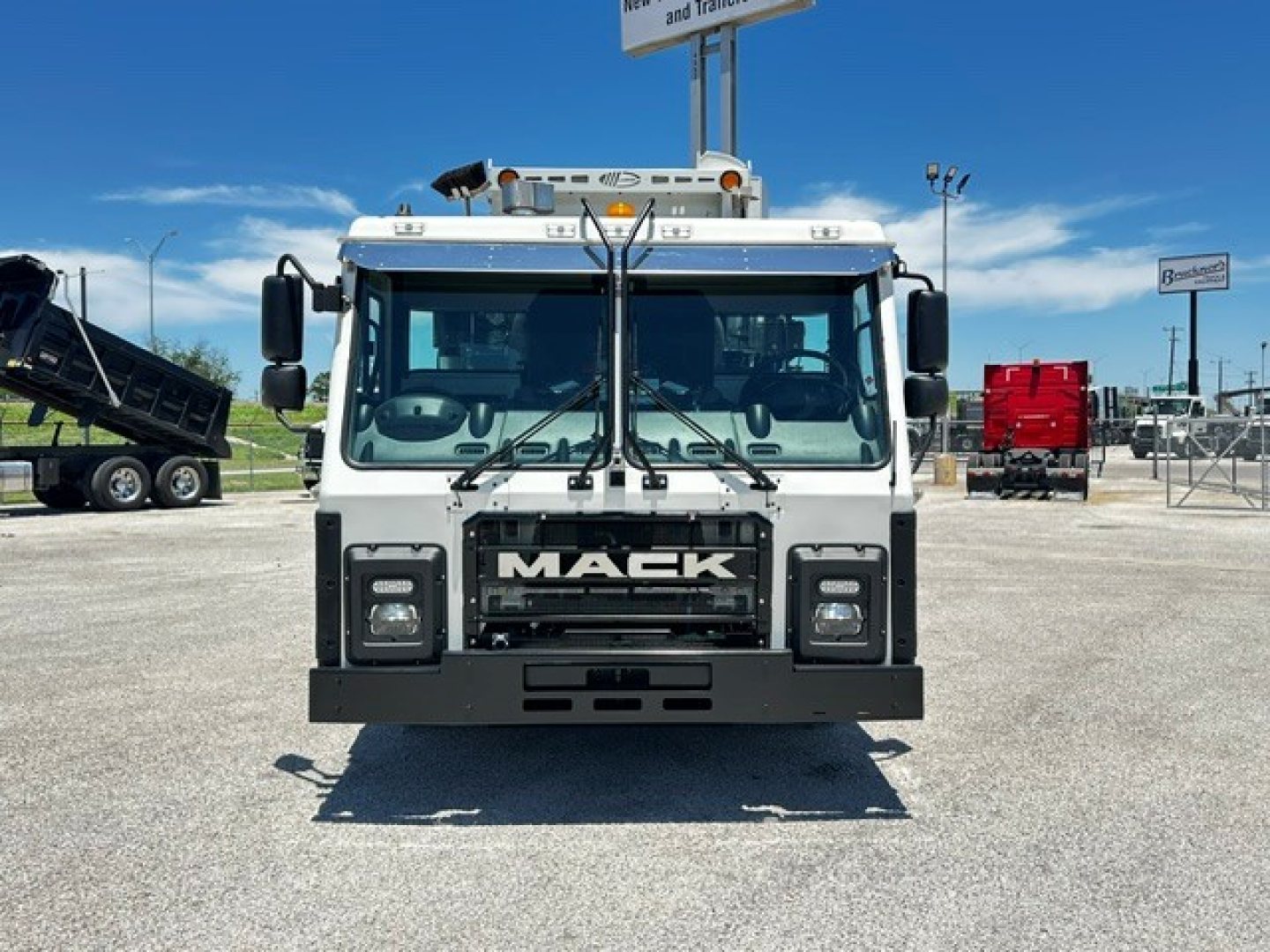 Front view of a white 2025 Mack LR64 truck showing grille with MACK lettering and bumper, with large windshield, mirrors, and rooftop lights.