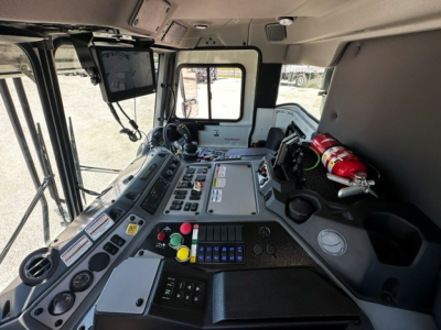 Truck cab interior showing control panel with numerous switches, gauges, and a mounted screen. Fire extinguisher secured on the right. Windows and mirrors visible.