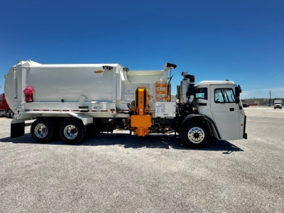 Mack LR64 refuse truck on level lot, side view showing large rear compactor body and orange hydraulic mechanism.