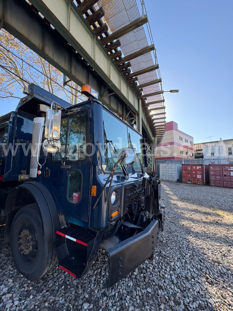 Front-left view of a blue Mack MRU600 dump truck in a yard with gravel, showing the cab and front equipment.