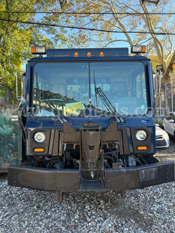 Front view of a 2010 Mack MRU600 dump truck showing chrome grill, headlights, and snow plow mounting; windshield wipers visible; side mirrors and roof light bar