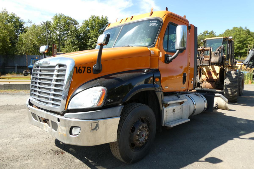 Truck cab close-up showing orange Freightliner Cascadia 125 with chrome bumper and front grille, VIN tag and stock number on frame.