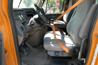 Truck cabin interior of a Freightliner Cascadia 125; driver seat with orange seatbelt, dashboard gauges, steering wheel, gray upholstery.