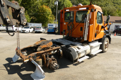 Orange Freightliner Cascadia 125 daycab truck with a decommissioned front axle and exposed drivetrain; cab in view with transmission area and side steps.