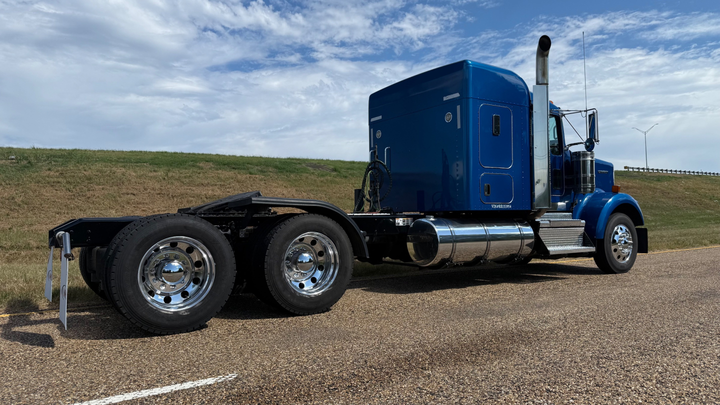 Kenworth W900 conventional sleeper truck side view showing blue cab and dual rear axles with chrome fuel tanks and exhaust stack.