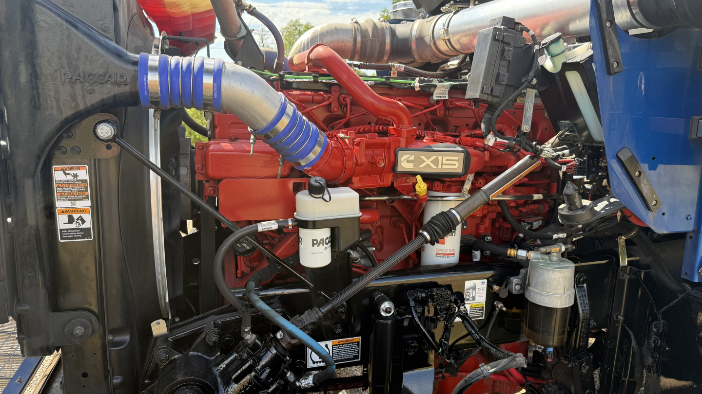 Engine bay of a 2024 Kenworth W900 showing CX15 engine with red block, blue hoses, white brake fluid reservoir, and various black hoses.