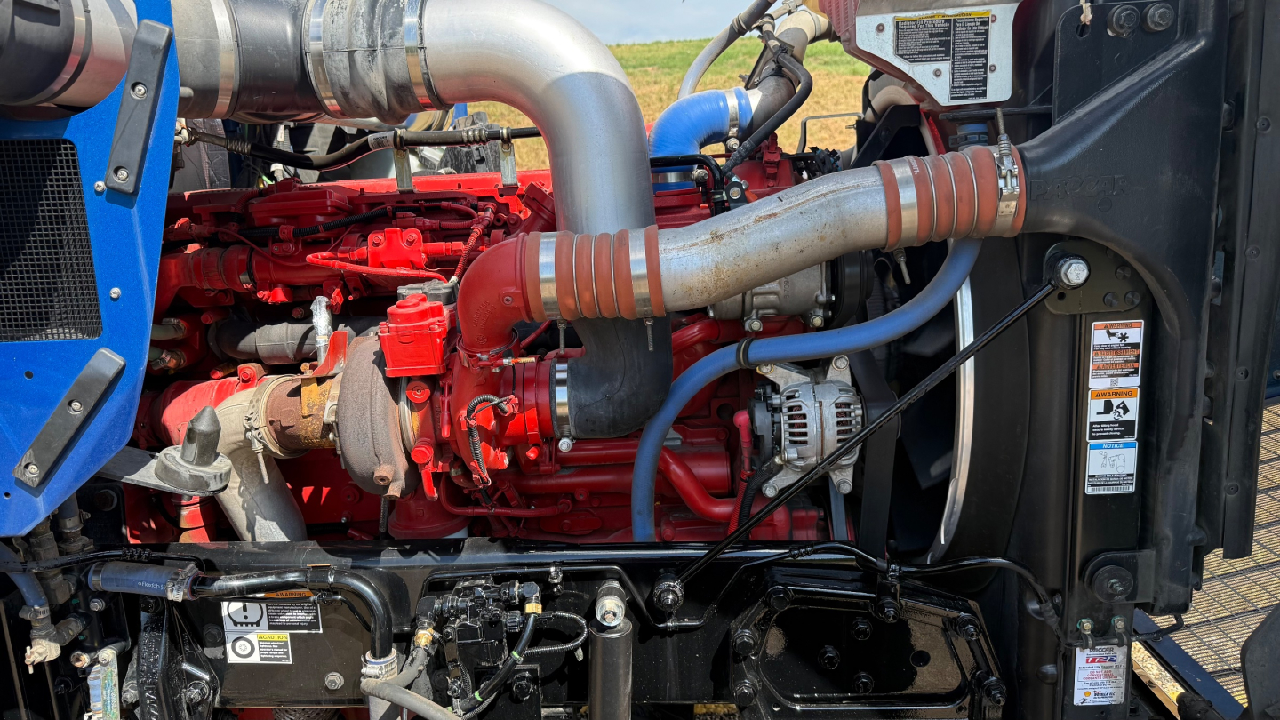 Kenworth W900 truck engine bay showing red engine block, silver exhaust manifold, blue hoses, and black radiator support. Visible labels and hoses.
