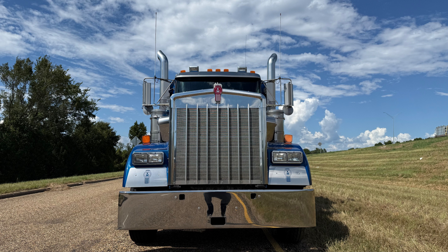 Front view of a blue Kenworth W900 truck with chrome grille and large bumper, daytime.