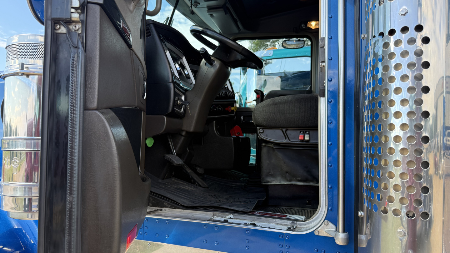 Kenworth W900 truck interior viewed from open driver door showing seat, steering wheel, and dashboard.