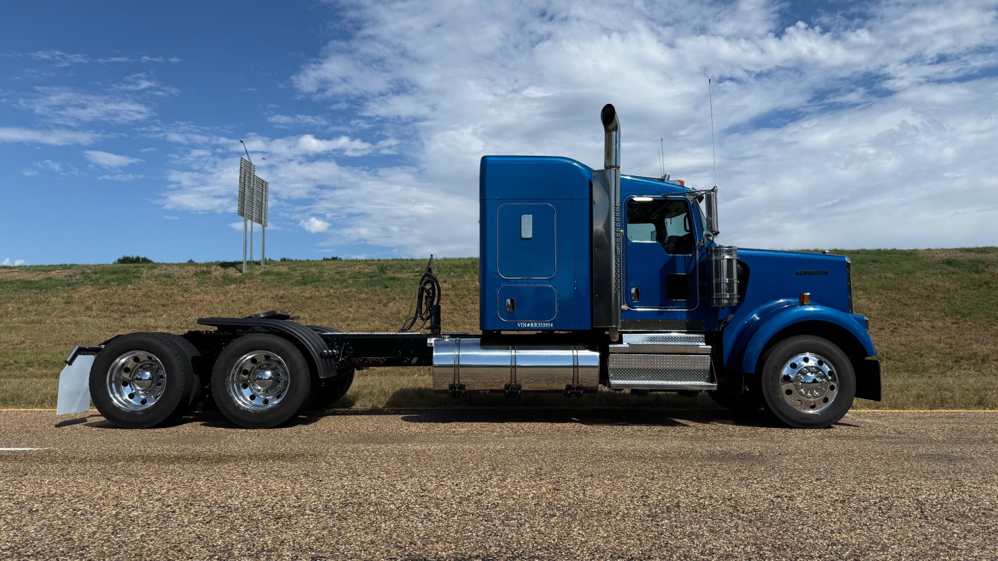 Side view of a blue 2024 Kenworth W900 conventional sleeper truck showing chassis, rear tandem axles, exhaust stack, and side fuel tanks.