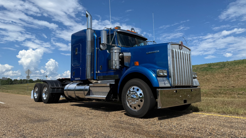 Truck cab exterior side view of a blue 2024 Kenworth W900 Conventional Sleeper with chrome accents and dual rear wheels.