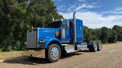 Kenworth W900 conventional sleeper truck in blue with chrome bumper and exhaust stacks, front grille, and dual rear axles on a road. Clear sky and trees in background.