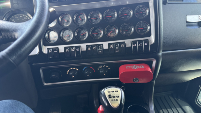 Kenworth W900 truck dashboard showing multiple gauges and control switches with red miscellaneous switch cover in center console.