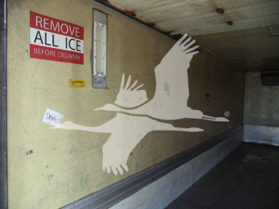 Interior of a refrigerated Mack MD6 truck; wall with a large white goose graphic, 'REMOVE ALL ICE BEFORE DELIVERY' sign, and a small window/vent.