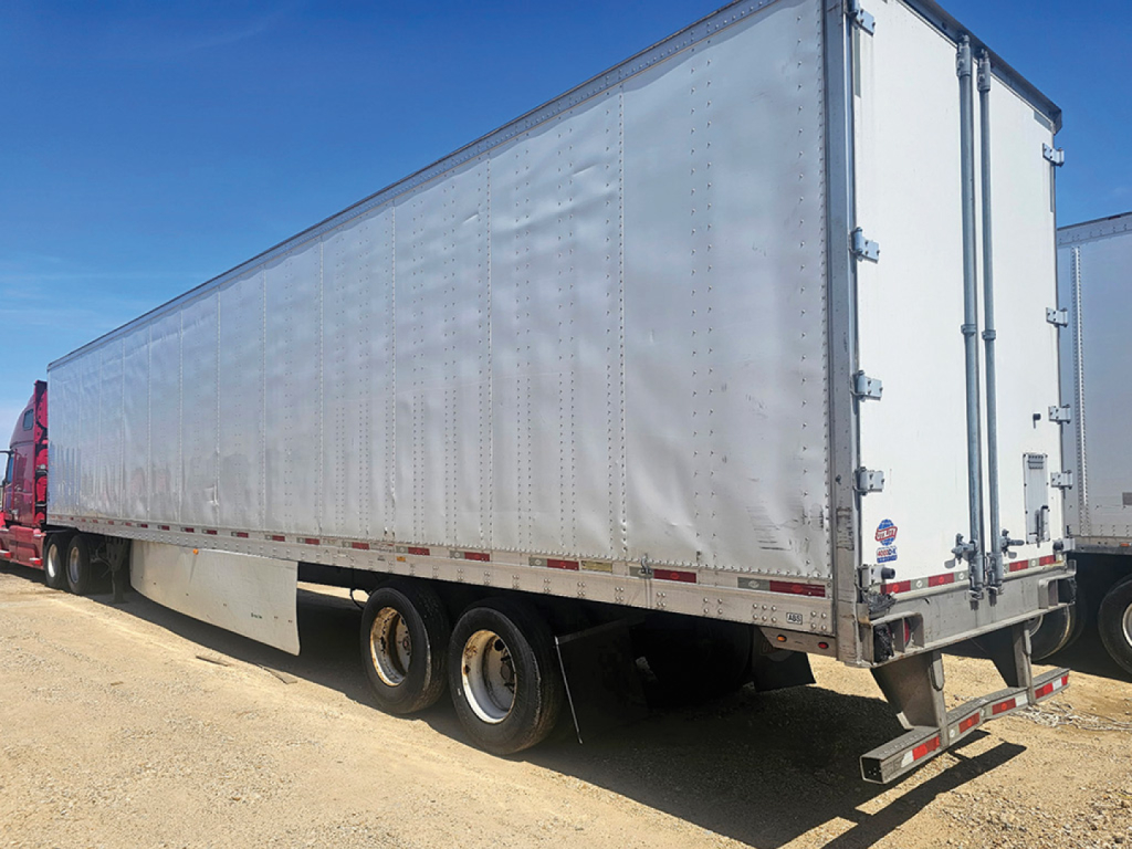 Long silver Utility trailer showing triple-axle rear wheels and rear doors with hinges and latches. Side and rear reflective tape. Clear sky.