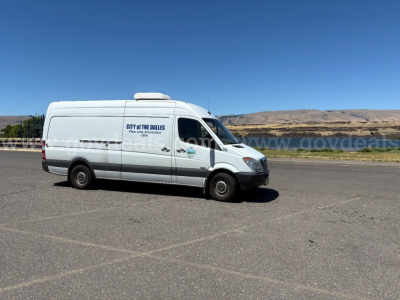 White Freightliner Sprinter cargo van with City of the Dalles logo on door, side windows, and black tires, parked on paved lot with hills in background.