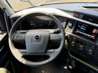 Volvo truck interior steering wheel and dashboard with digital gauge cluster and touchscreen infotainment system; visible text on screen and buttons.