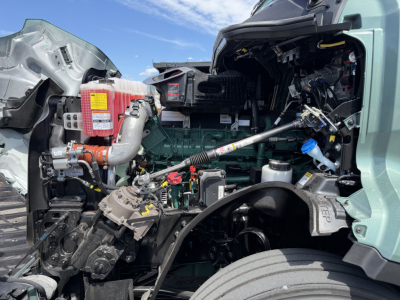 Volvo VNL truck engine bay as viewed from the driver's side with exposed components, hoses, and a yellow warning label on a red coolant reservoir.
