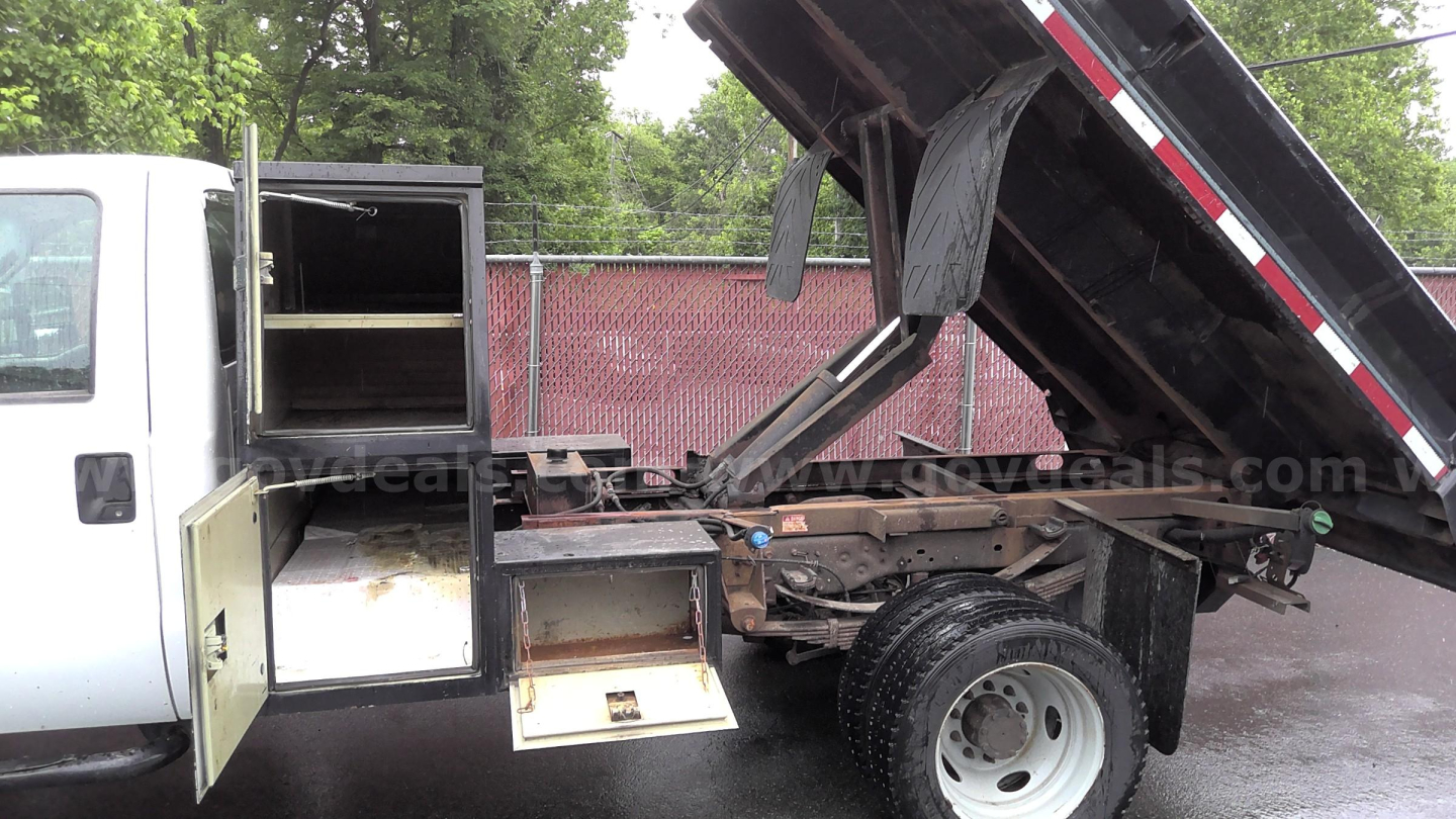 Ford F-450 dumping truck bed raised showing side storage cabinet and steps with toolbox compartment open.