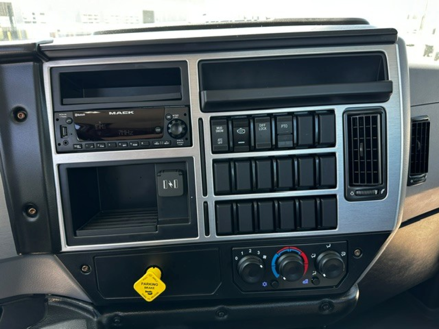 Mack MD6 cab dashboard showing steering audio system with 'MACK' badge, multiple switches, climate controls, and a yellow parking brake cap.