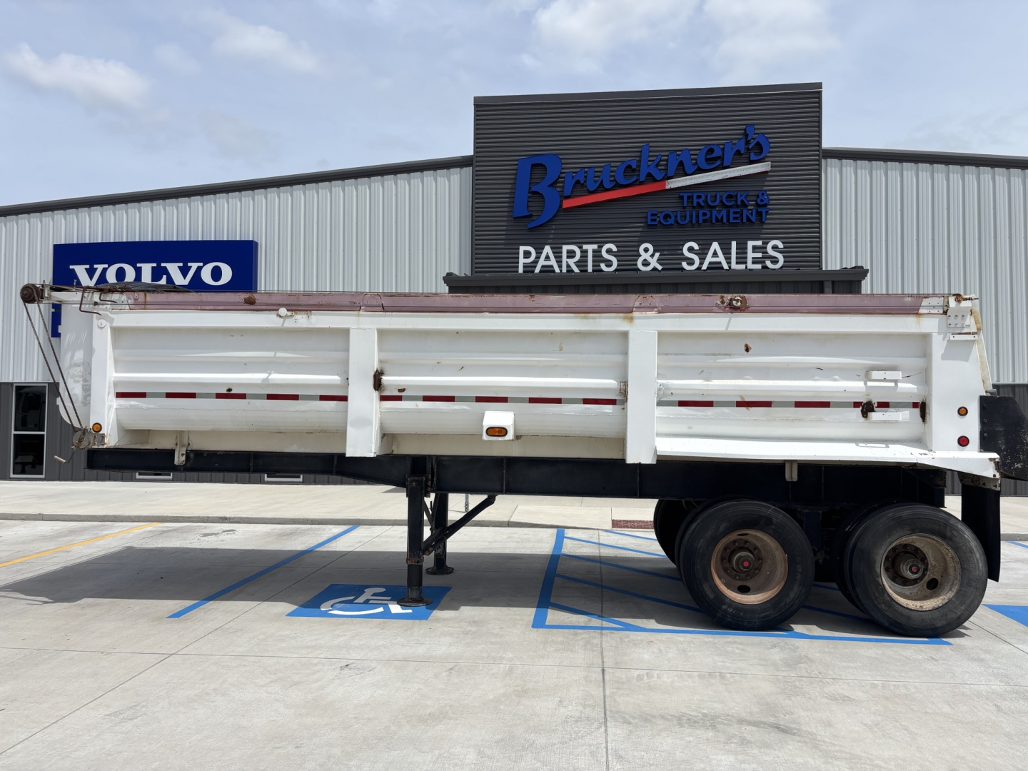 Trailer with white side panels and dual rear axles in front of a parts and sales building; visible signage includes Volvo on a blue sign and a Brackner? Truck & Equipment Parts & Sales sign.