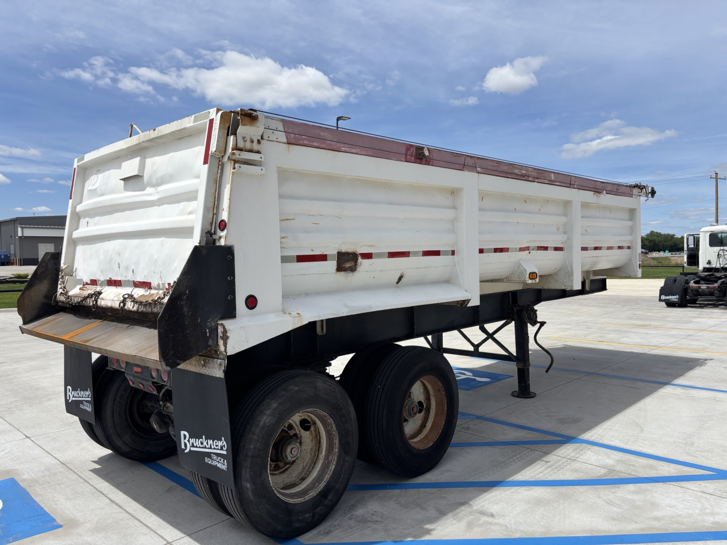 Dump trailer showing wheel wells and side panels with red reflective tape and platform tailgate, underclear sky.