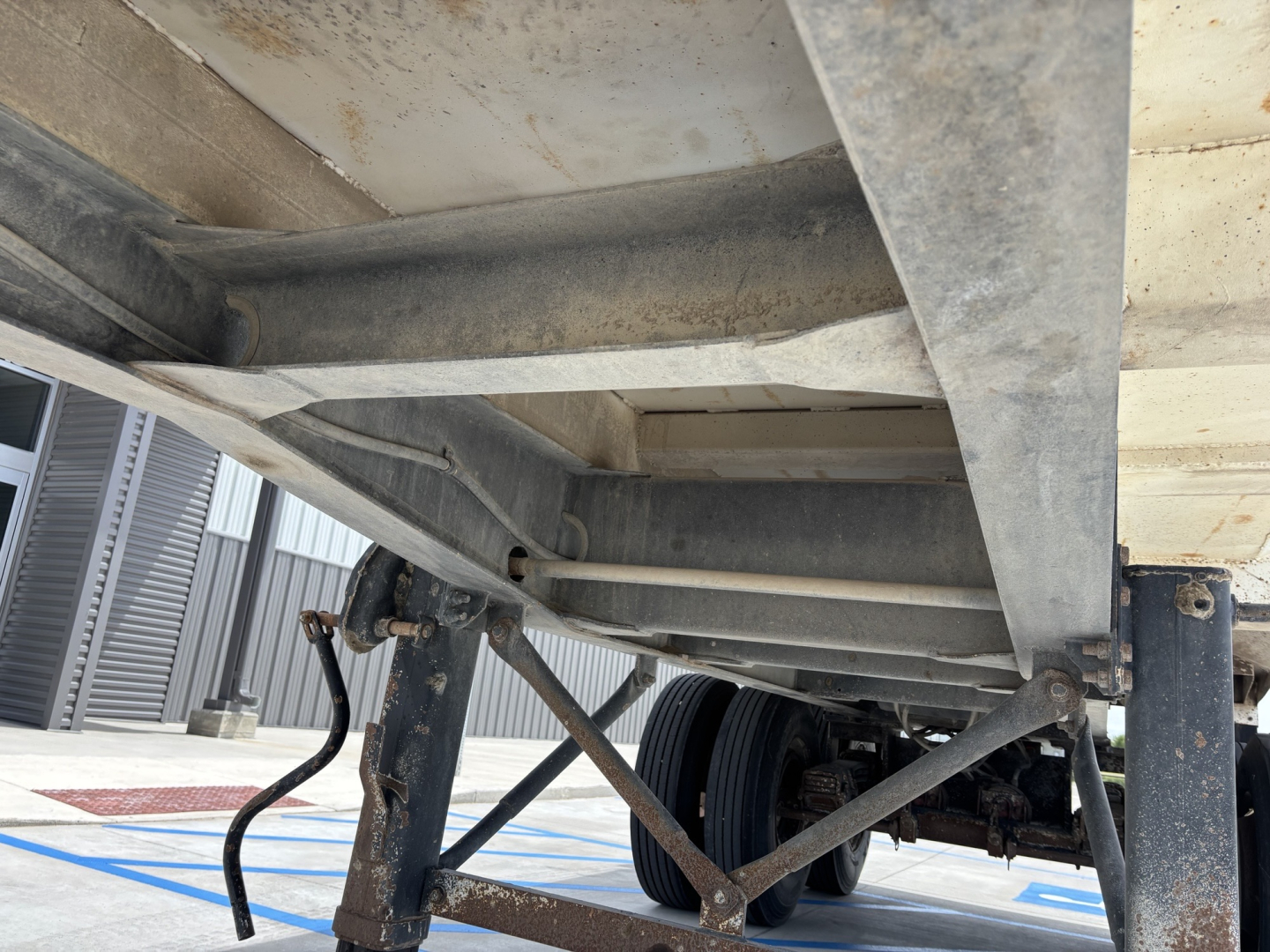 Dump trailer undercarriage showing frame rails, axles, leaf springs, and wheel assembly with visible metal components and weathering.