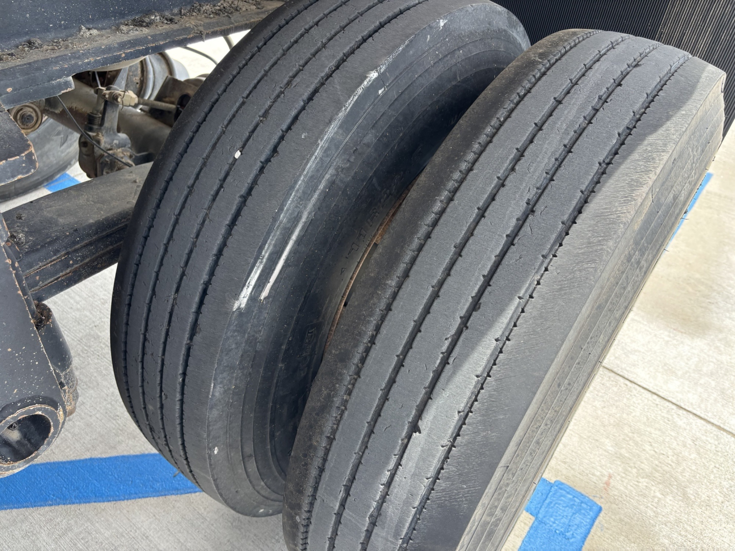 Dual rear wheels of a trailer with worn tread showing six straight grooves; part of chassis and suspension visible.