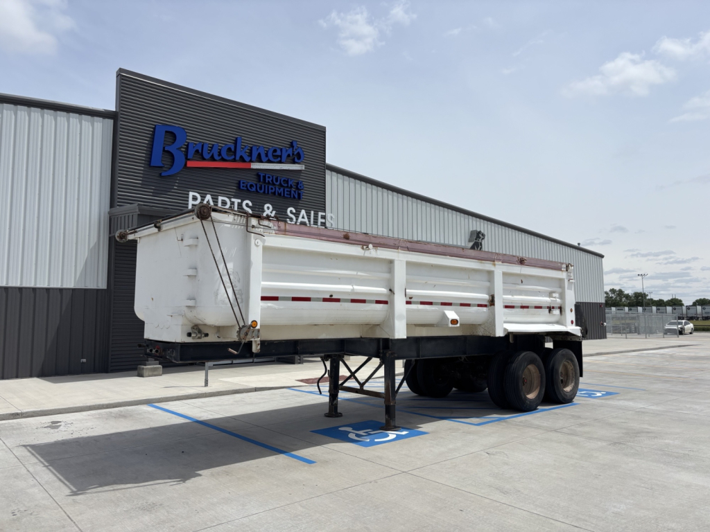 Dump trailer in front of a dealership building showing white steel body and dual rear axles with support legs extended.