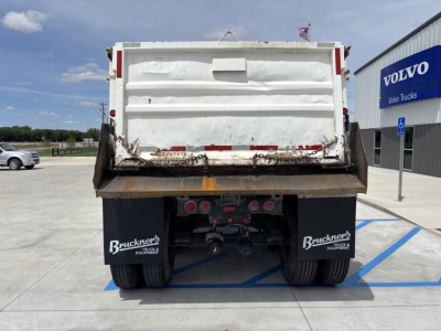 Rear view of a dump trailer with white tailgate, rusted bed edge, and dual rear axles. Bruckner's Equipment logos on mud flaps. Volvo dealership building in background.