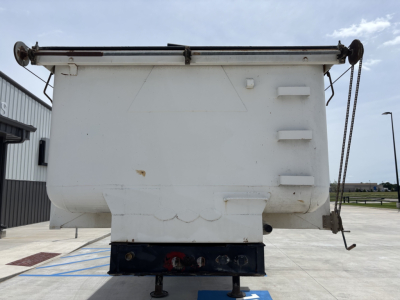 Dump trailer rear view showing spill guard and hydraulic tailgate with hinge pins, white body, black undercarriage, and chains at the right side.