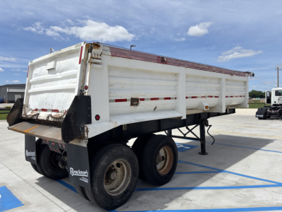 Dump trailer showing wheel wells and side panels with red reflective tape and platform tailgate, underclear sky.