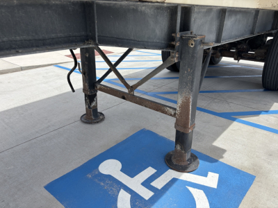 Trailer landing gear legs and frame support on a dump trailer, with blue accessible parking marking visible on the ground.
