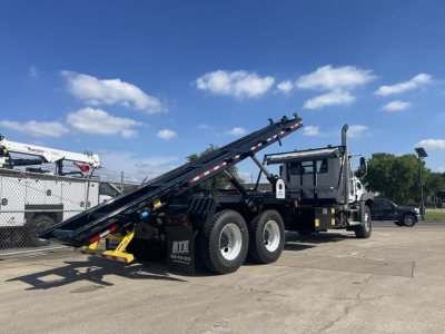 Mack GR64B rolloff system on a new truck with hydraulic bed extended, dual rear axles, cab with mirrors, VIN 1M2GR2GC8VM058627 visible listing details not on image.