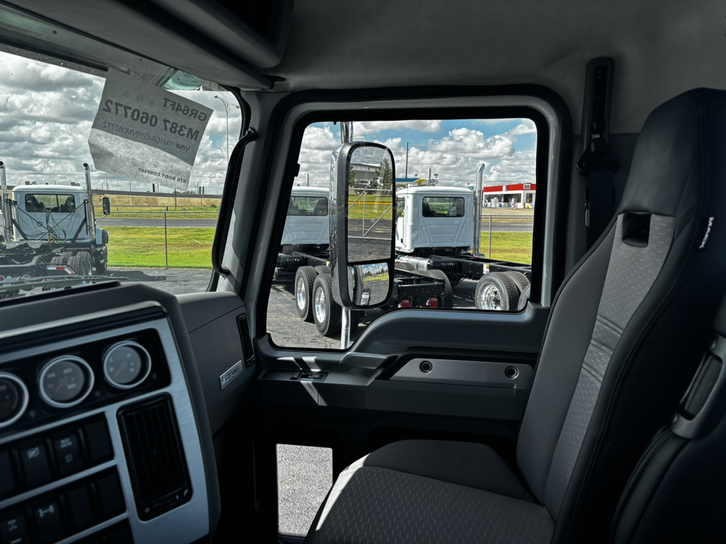 Truck cab interior view of a 2027 Mack GR64FT showing steering wheel, dashboard gauges, and passenger seat. Exterior side mirror and other trucks visible through window.