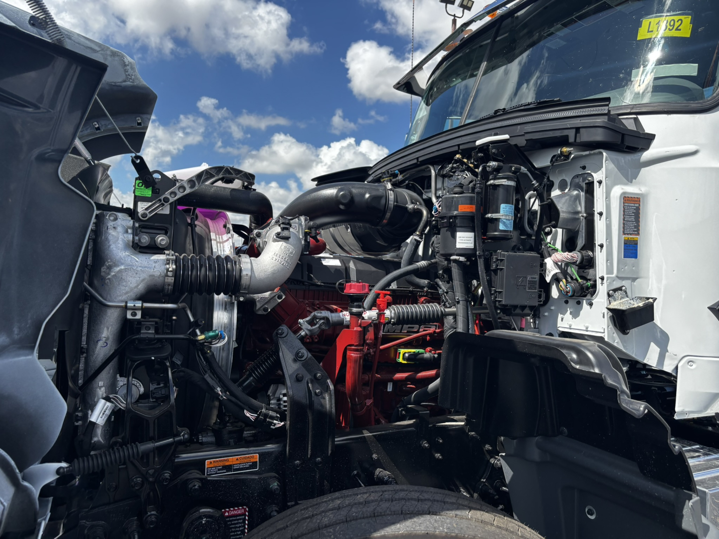 Truck engine bay of a 2027 Mack GR64FT visible with exposed components, hoses, and battery visible; front cab area and hood open.