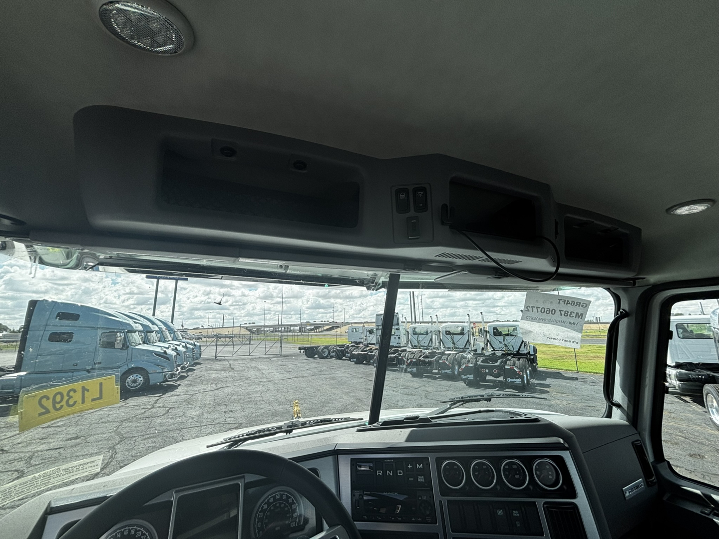Interior cab view of a 2027 Mack GR64FT from driver’s seat showing dashboard gauges, steering wheel, overhead module, and windshield with other trucks outside.